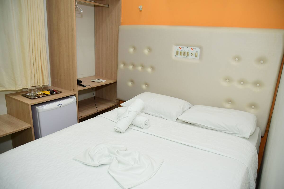 A hotel room with a bed, a small refrigerator, a desk with a lamp, and a power outlet on the wall.