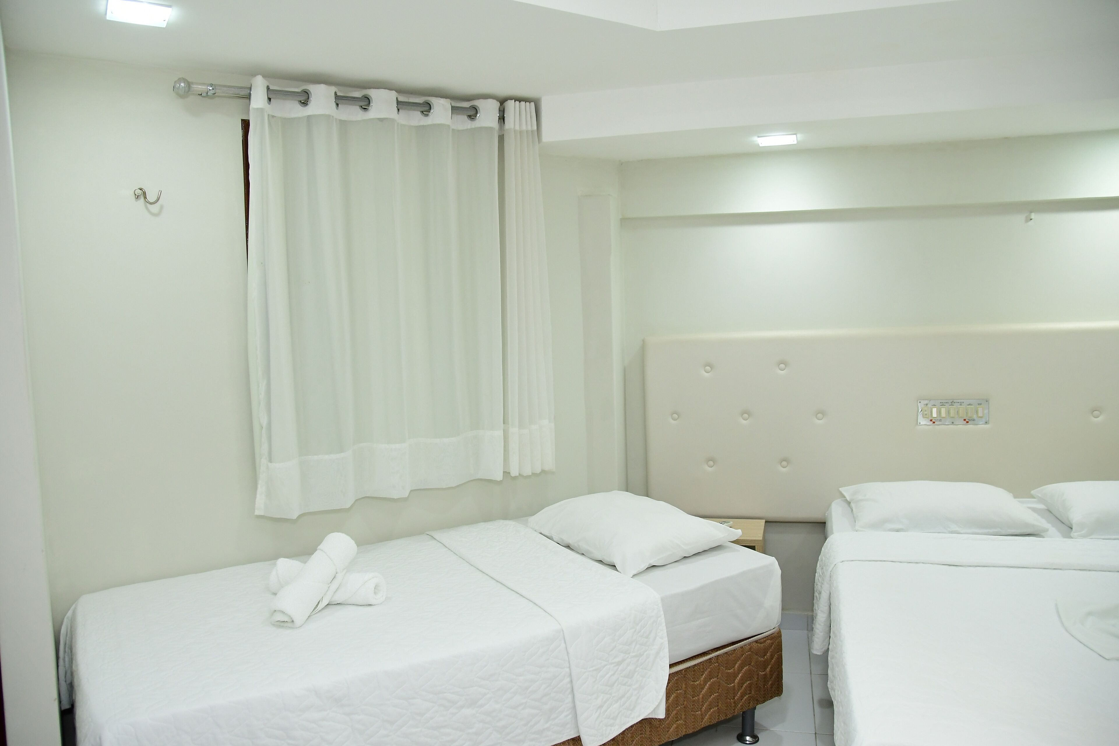 A hotel room with two single beds, a large window with white curtains, a wall-mounted light fixture, and a power outlet on the wall.