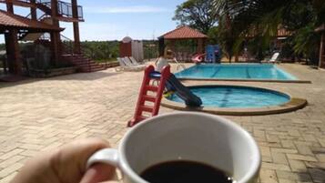 A hand holding a cup of coffee with a pool and lounge chairs in the background.
