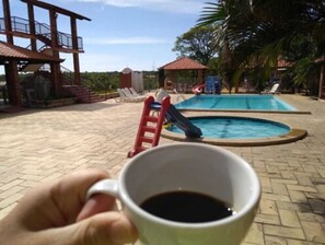 A hand holding a cup of coffee with a pool and lounge chairs in the background. - Hotel Serrano (Martins)