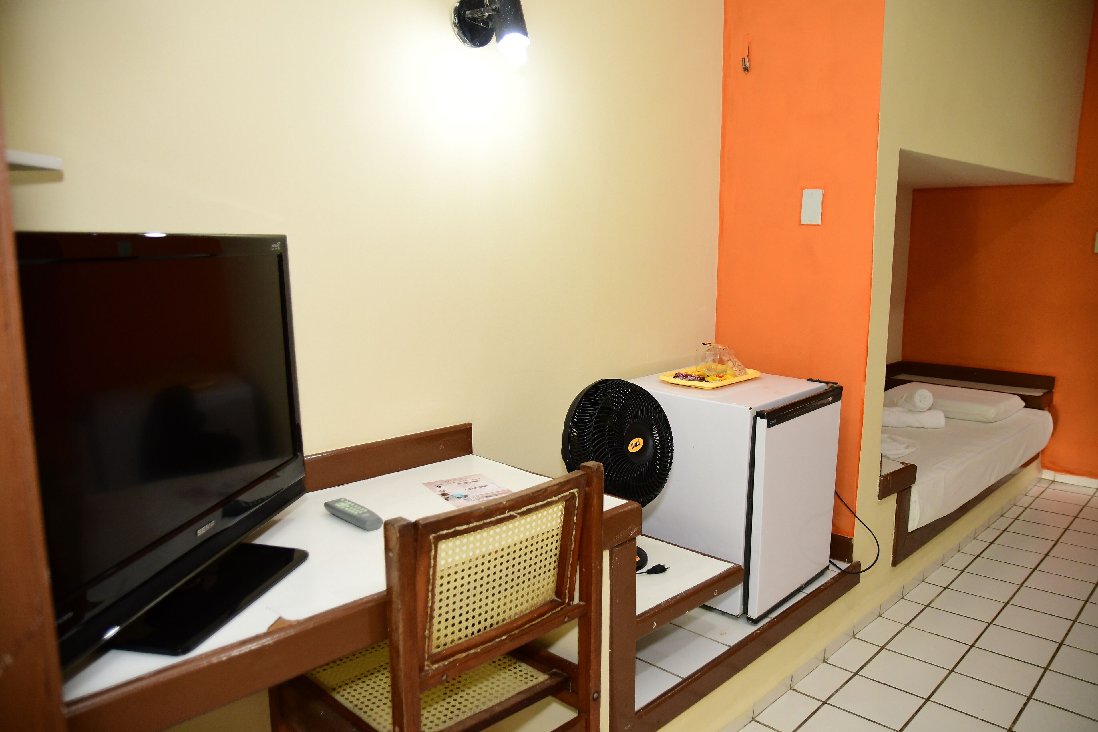 Una habitación pequeña con televisión, un refrigerador, un ventilador y una cama.