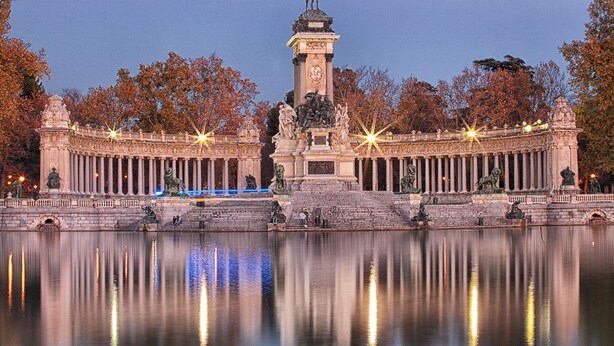 A large, historic building with a central statue, surrounded by a reflective water body and trees with autumn foliage. - La Reina de Gran Via (Madrid)