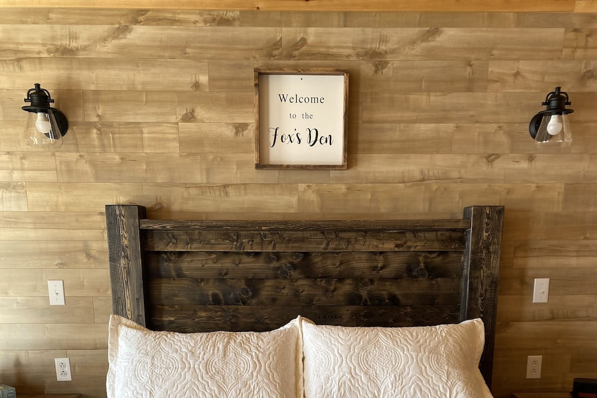 Un lit avec une couette blanche et un bordure rouge, une tête de lit en bois et un panneau d’accueil accroché au mur.