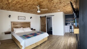 A bedroom with a wooden ceiling, a bed with a colorful quilt, a ceiling fan, and a view into the kitchen.