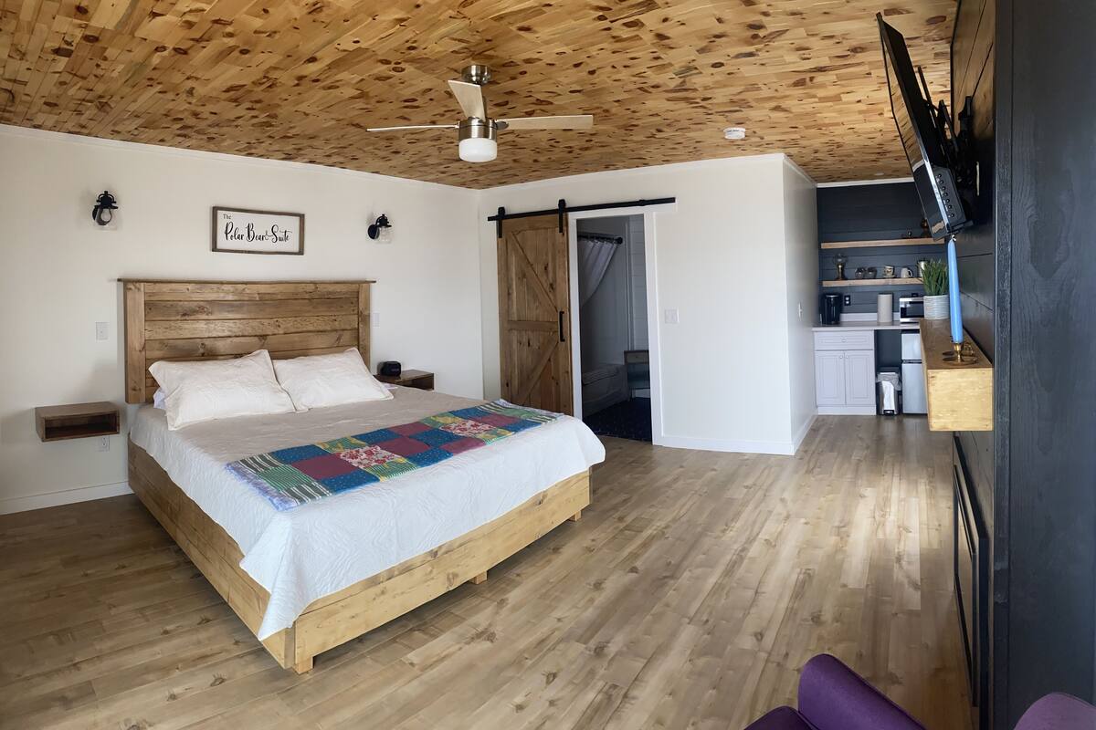 Une chambre avec un plafond en bois, un lit recouvert d’une couette aux couleurs vives, un ventilateur de plafond et une vue sur la cuisine.