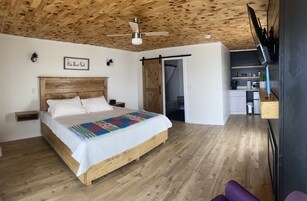 Un dormitorio con techo de madera, una cama con colcha colorida, un ventilador de techo y vista a la cocina.