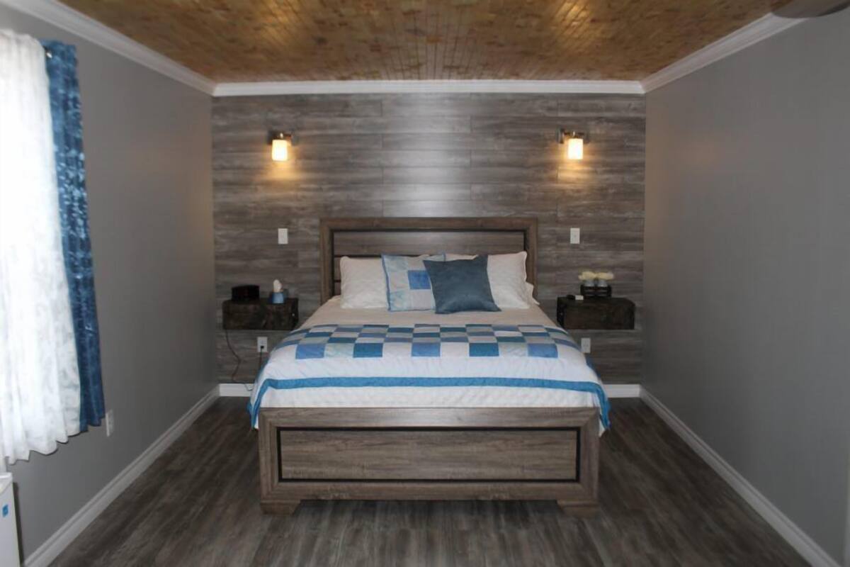 Une chambre à coucher avec une tête de lit en bois, une couvre-lit à carreaux, deux tables de chevet et des lampes fixées au mur.