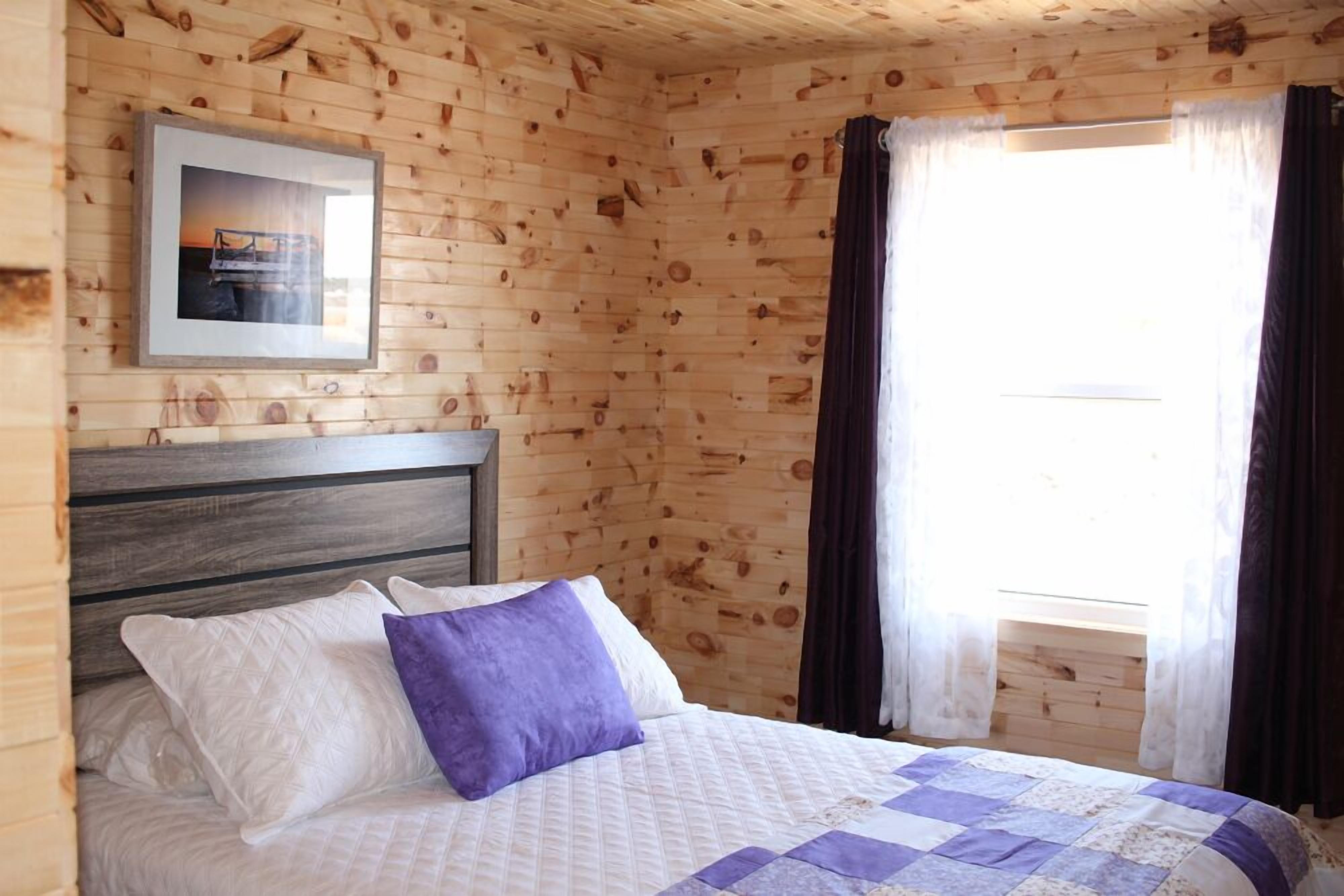 A wooden cabin room with a bed, a framed picture on the wall, and a window with curtains.