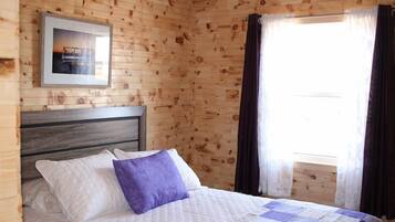 A wooden cabin room with a bed, a framed picture on the wall, and a window with curtains.