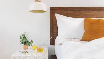 Ein ordentlich bezogenes Bett mit weißen und senffarbenen Kissen, ein kleiner Nachttisch mit Pflanze, einem Glas Wasser und einer Lampe.