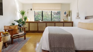 A bedroom with a large bed, a wooden nightstand, a window with a view of greenery, and a bedside table with a lamp. - Silver Lake Pool & Inn (Los Angeles)
