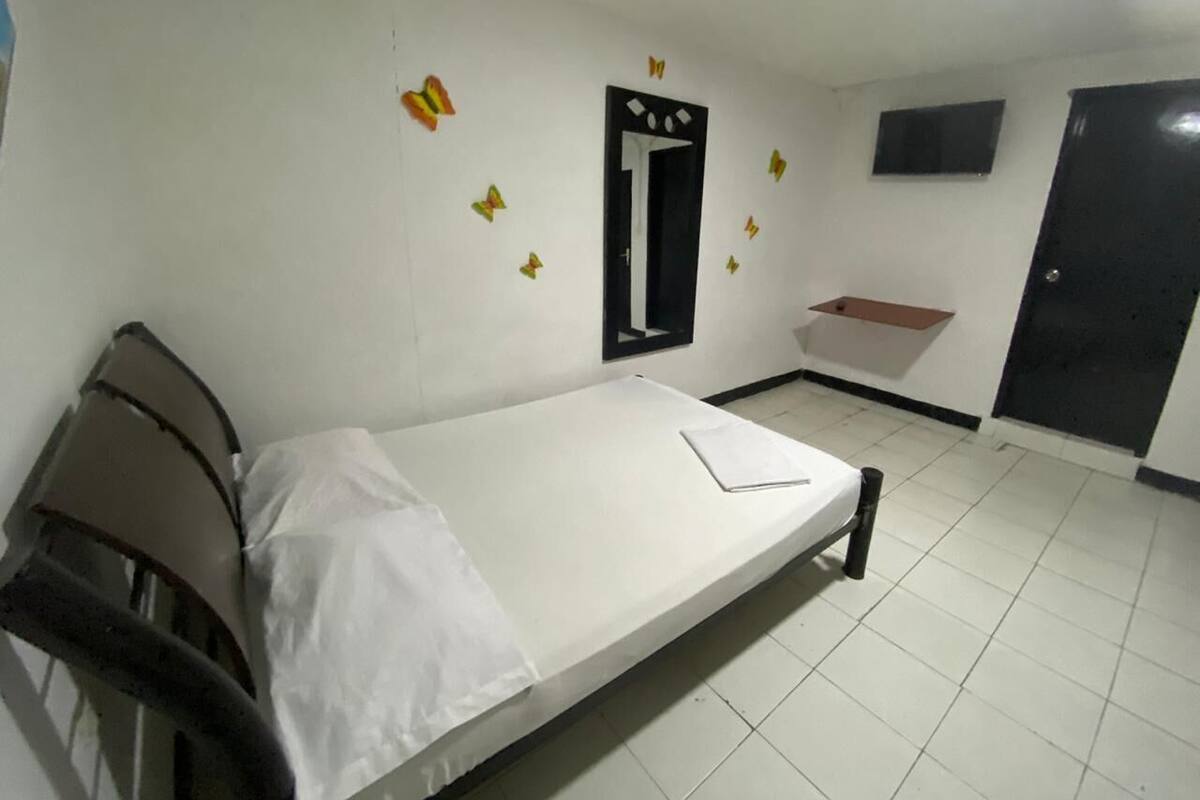 A single-bed room with a white mattress, a wooden headboard, a small bedside table, a mirror, a TV mounted on the wall, and a door with a black frame.