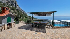A stone patio with a table and chairs, a small brick structure with a green door, and a pool area with a view of the sea. - Villa Makar with  infinity heated pool,gym,grill, 3 bedrooms,3 bathrooms (Dalmatia)