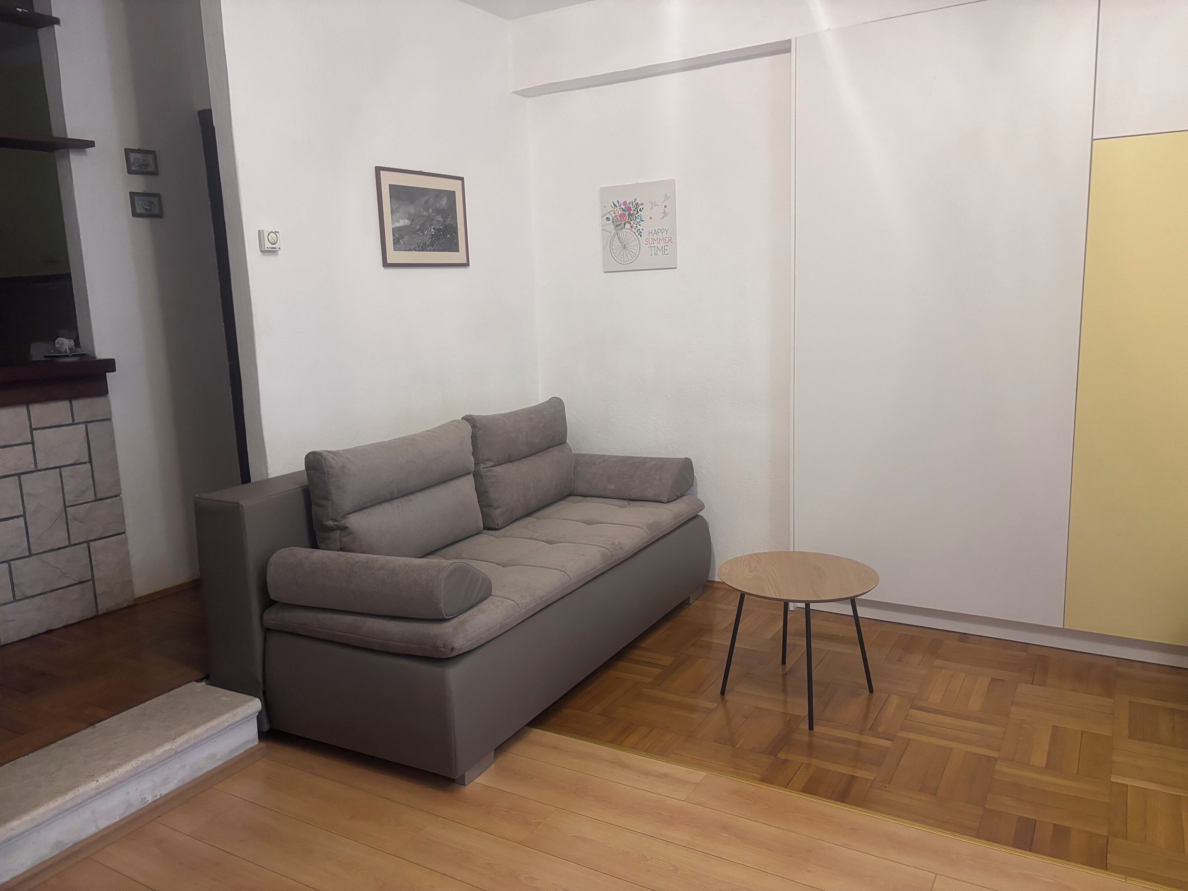 A modern living room with a grey sofa, a small round wooden table, and a herringbone-patterned wooden floor.