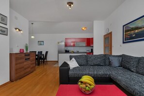 A modern living room with a grey sofa, a dining area with a wooden cabinet and chairs, and a kitchen with red cabinets and a white countertop. - Villa Makar with  infinity heated pool,gym,grill, 3 bedrooms,3 bathrooms (Dalmatia)