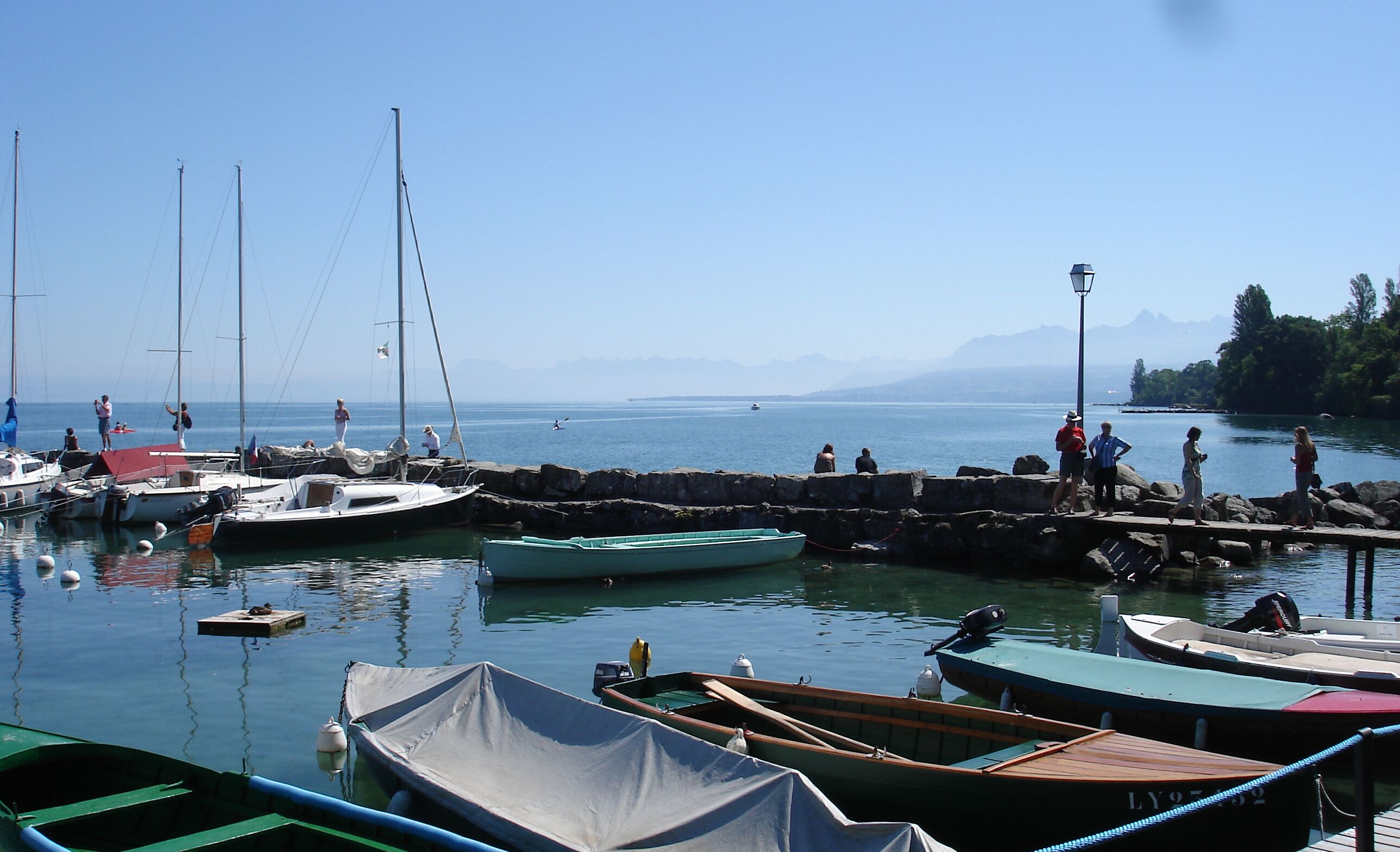 Un port de plaisance où sont amarrés différents types de bateaux, une jetée en pierre et des personnes qui se promènent le long de la jetée.