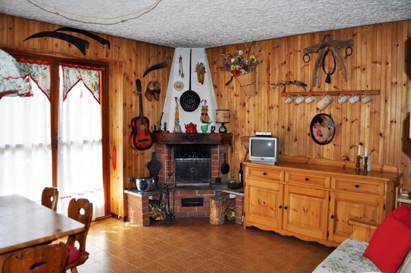 A cozy living room with a fireplace, wooden walls, a television, and a dining area with a table and chairs. - Rustic in the Chapelle complex (Sauze d` Oulx)