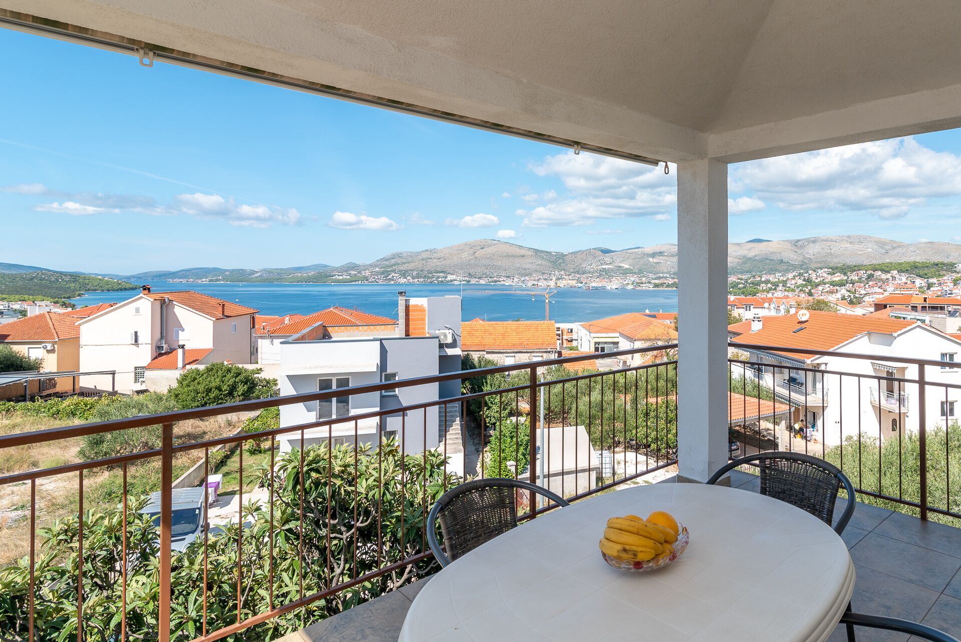 A balcony with a table and chairs, overlooking a coastal town with red-roofed buildings and a view of the sea.