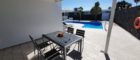 A covered patio with a table and chairs, a swimming pool in the background, and a stone wall with a lifebuoy.