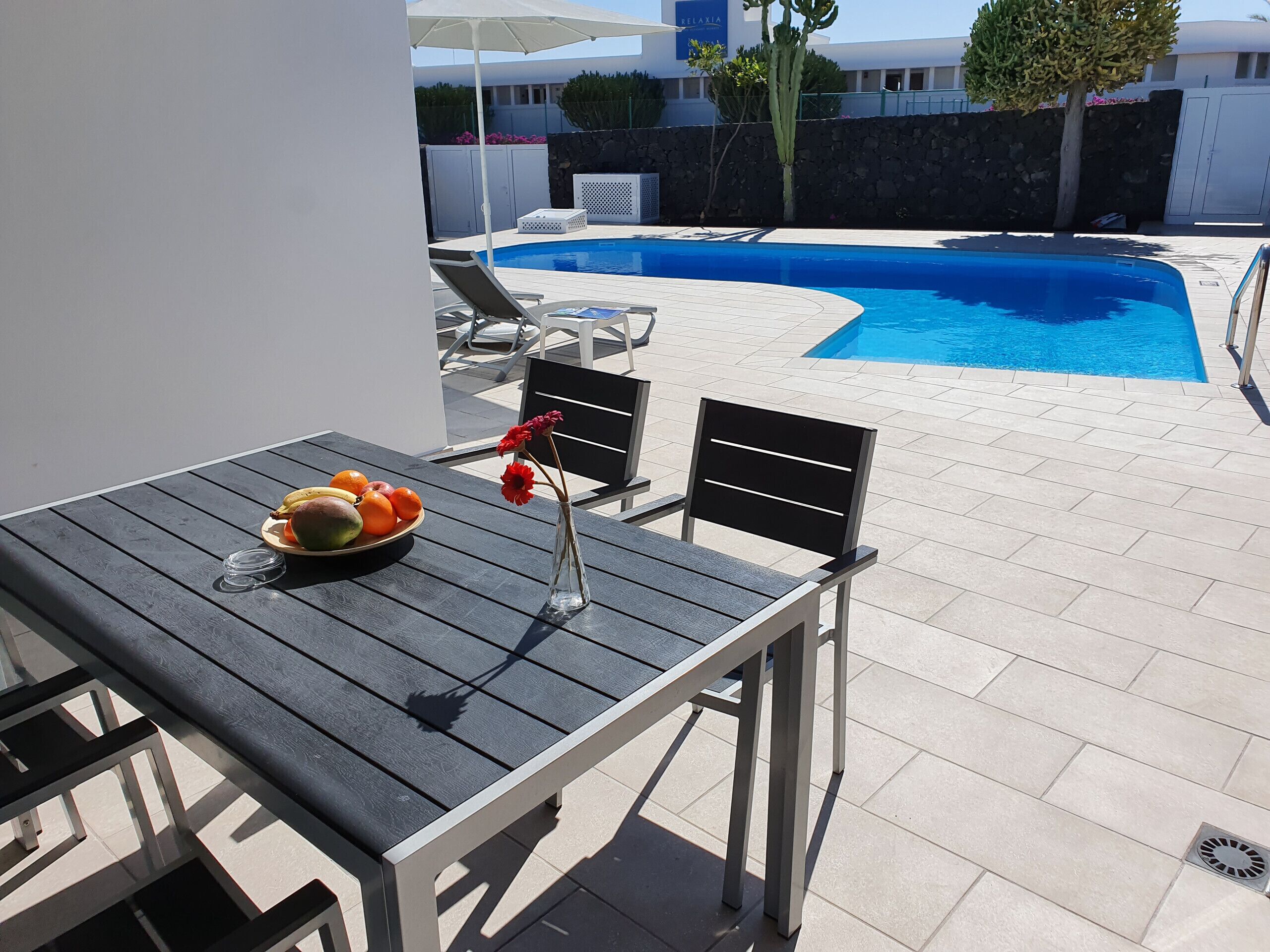 Ein Tisch am Pool mit Obstschale und Blumen, umgeben von Liegestühlen und einem Schwimmbecken.