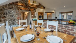 A dining area with a wooden table set for four, surrounded by white chairs. A brick wall and a stone archway are visible in the background. - Mayes Cottage, Brancaster, Norfolk (Brancaster)