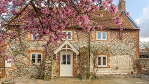 A stone house with a white door and windows, surrounded by gravel and flowering trees. - Mayes Cottage, Brancaster, Norfolk (Brancaster)