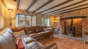 A cozy living room with a leather sofa, a fireplace, and exposed wooden beams. - Mayes Cottage, Brancaster, Norfolk (Brancaster)