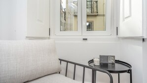 Una stanza con un divano bianco, un piccolo tavolino con una radio e una finestra che dà su un edificio con balconi.
