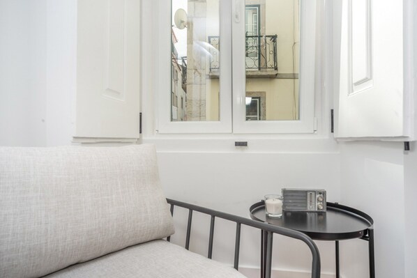 Una habitación con un sofá blanco, una mesita redonda con una radio y una ventana que da a un edificio con balcones.