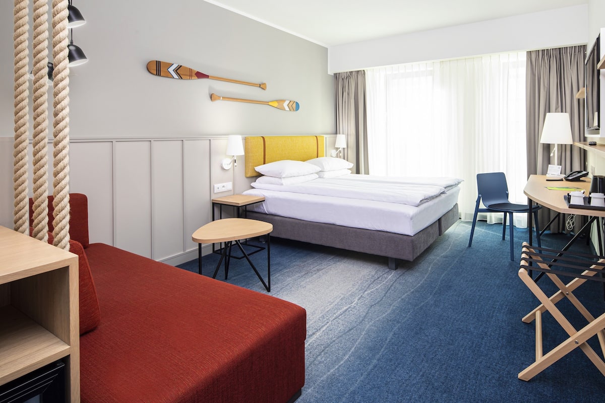 A hotel room with a bed, a desk, a chair, a small table, and a red sofa.