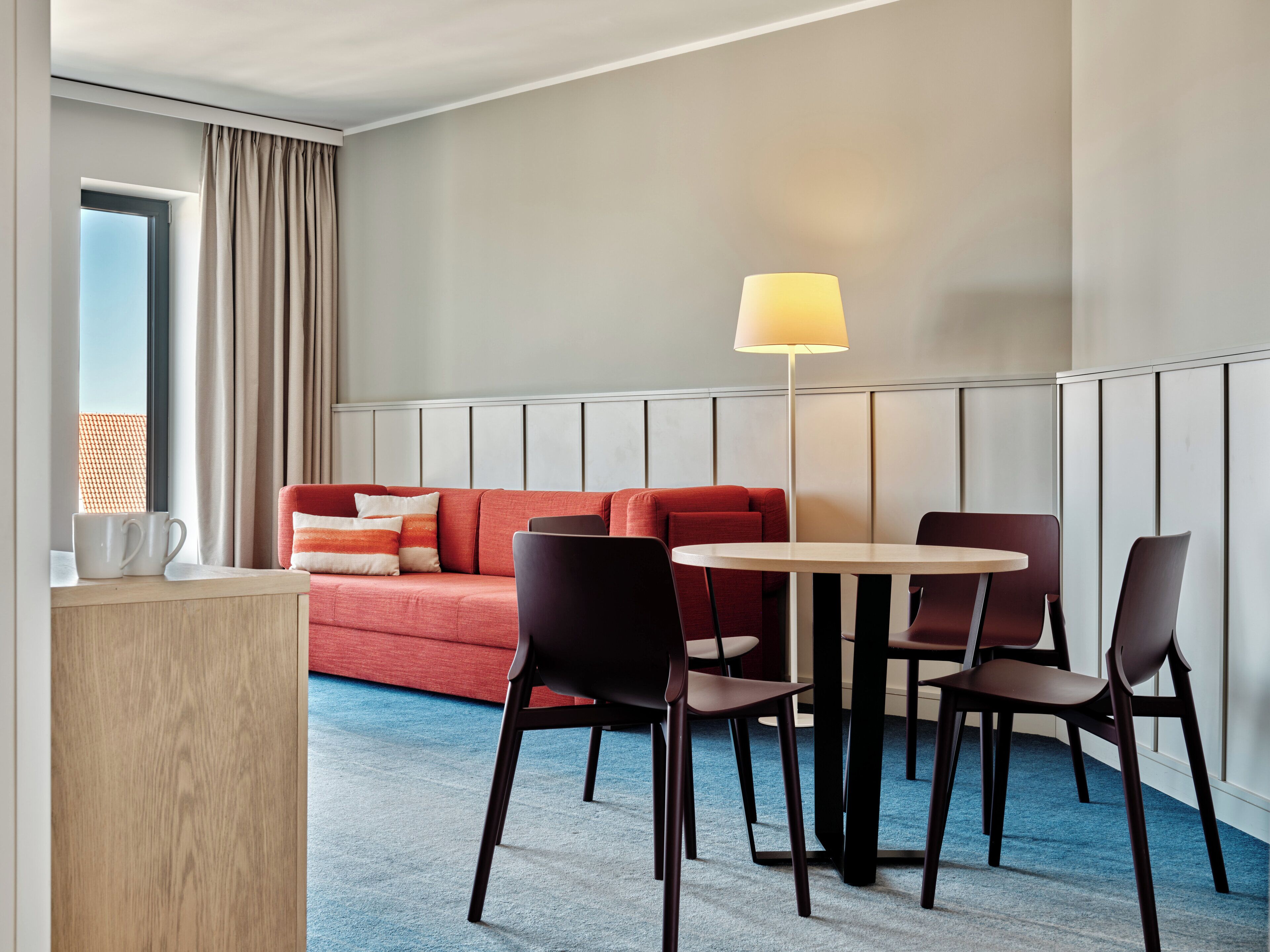 Habitación de hotel con un sofá rojo, dos sillones, una mesita, una lámpara y una ventana con cortinas.