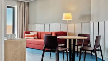 Habitación de hotel con un sofá rojo, dos sillones, una mesita, una lámpara y una ventana con cortinas.