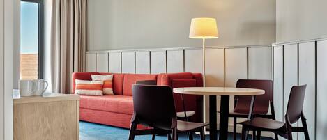 A hotel room with a red sofa, two chairs, a small table, a lamp, and a window with curtains.