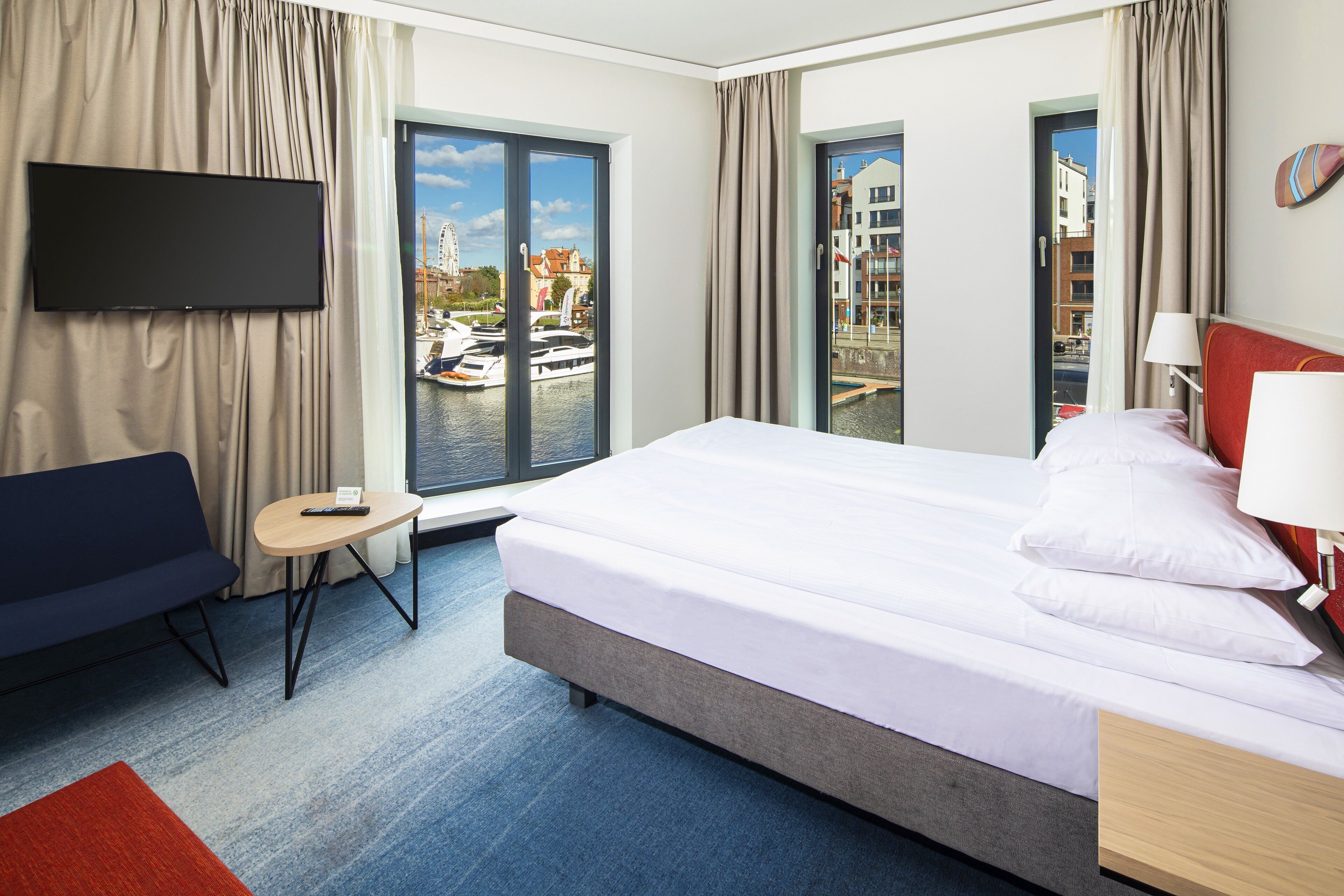 A hotel room with a bed, a TV mounted on the wall, a small table, and a view of a marina through the windows.