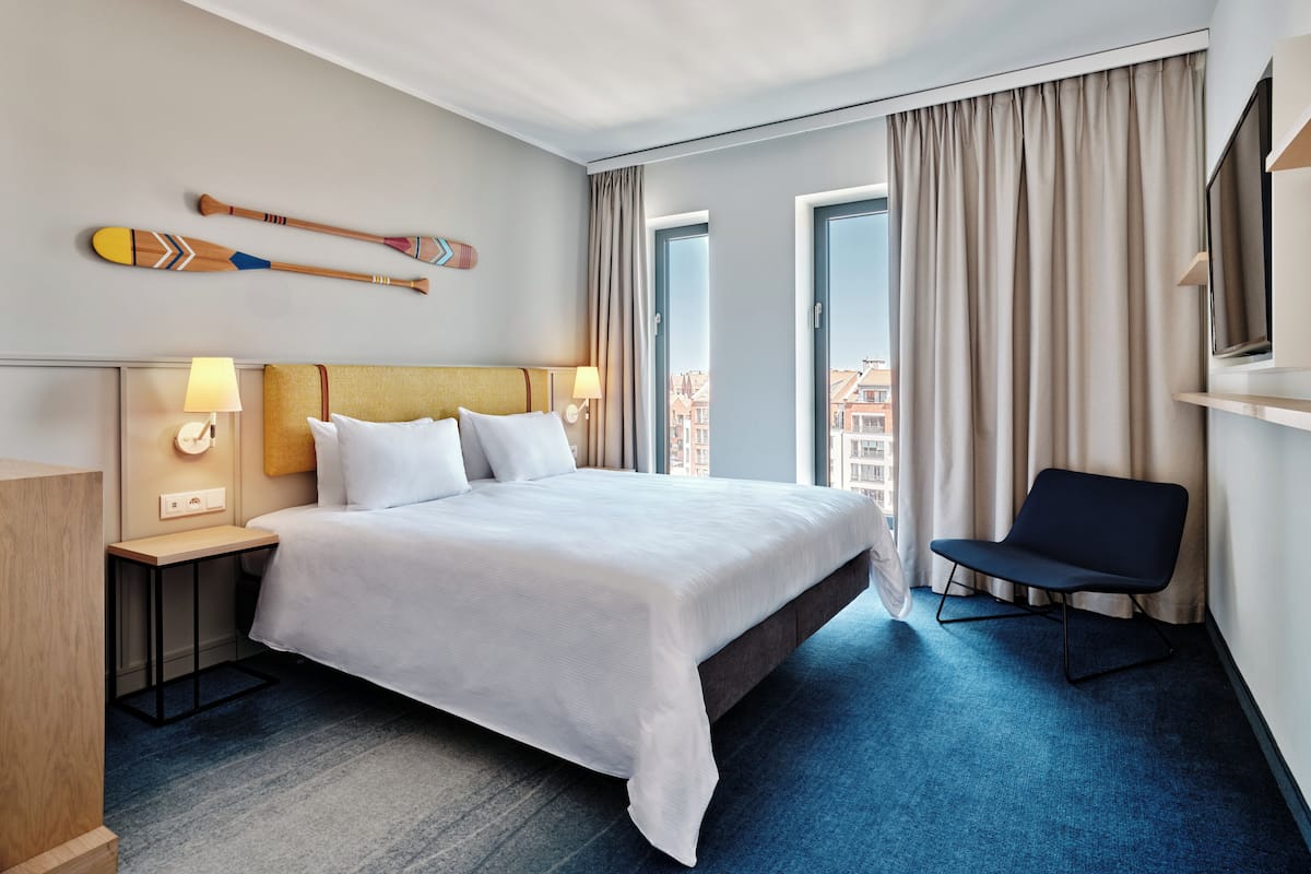 A hotel room with a large bed, a chair, a TV, and a view of the city through the window.