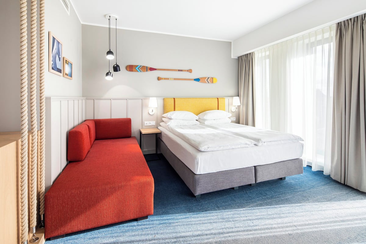 A hotel room with a bed, a red sofa, a nightstand, and a wall-mounted lamp.