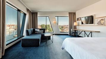 A modern hotel room with a large bed, a sofa, a desk, and a view of the city.