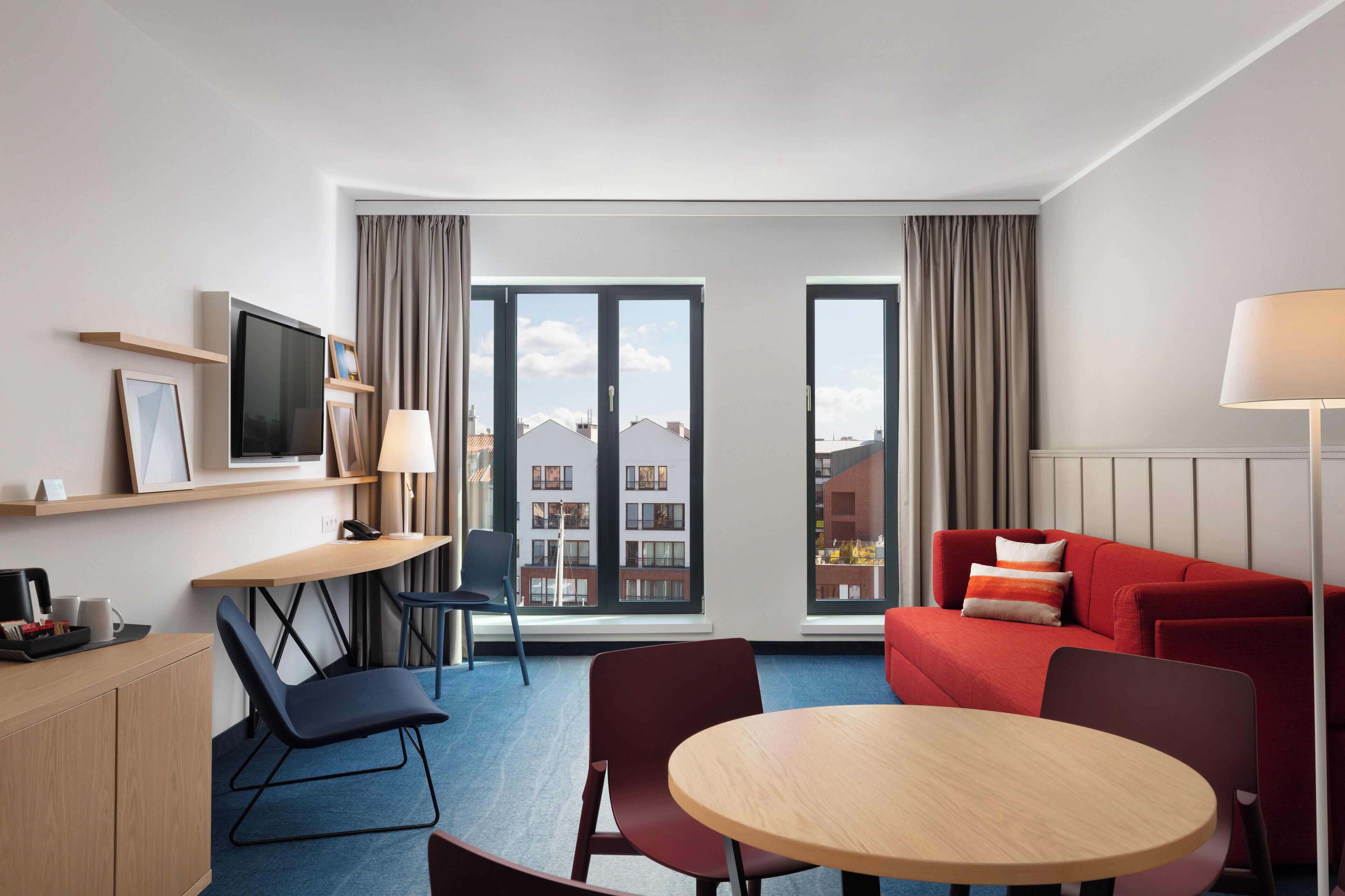 A modern hotel room with a desk, chair, round table, sofa, and large windows offering a city view.