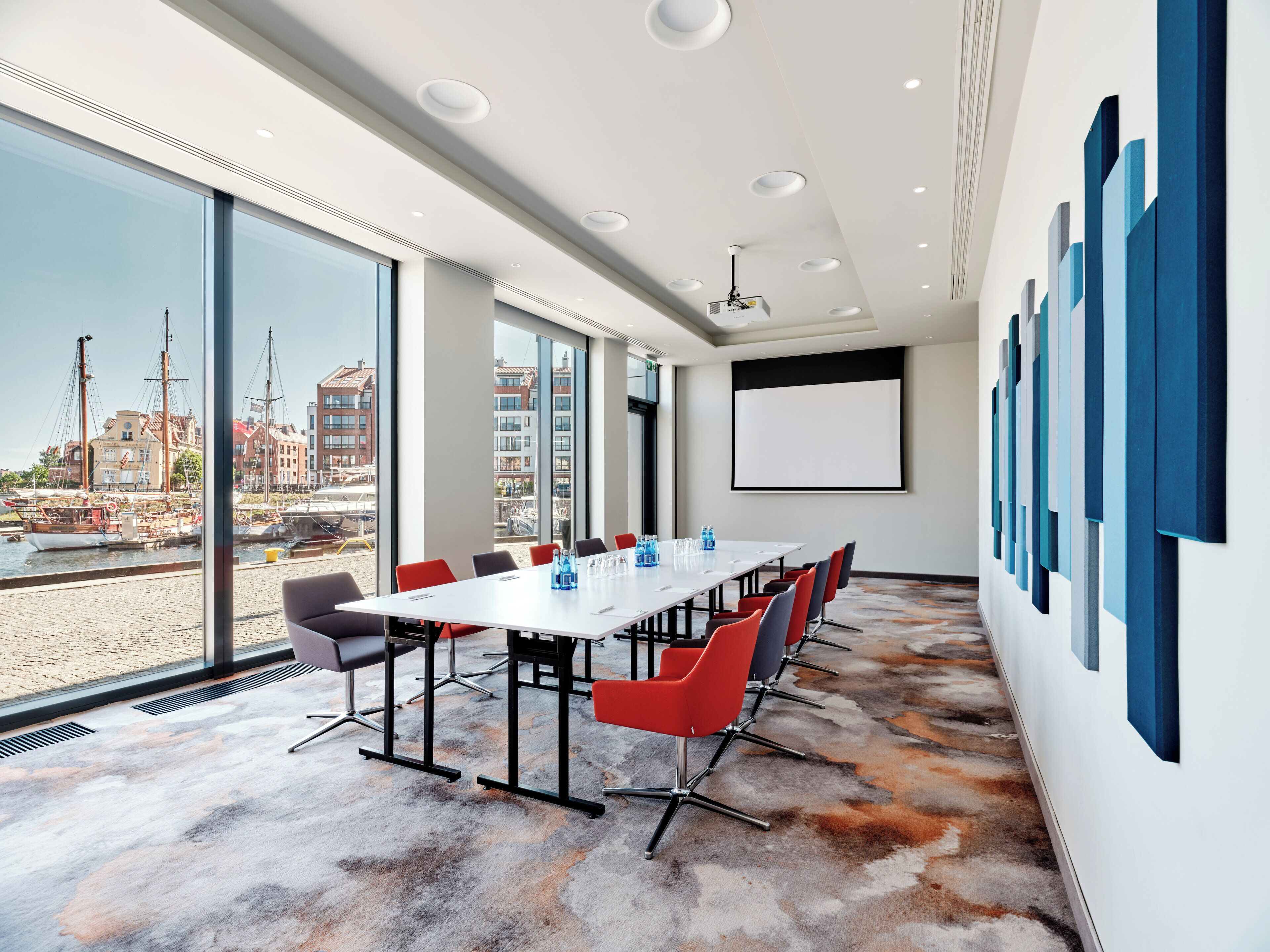 Una sala conferenze con un grande tavolo, sedie rosse e nere, uno schermo per proiettore e ampie finestre con vista sul porto.