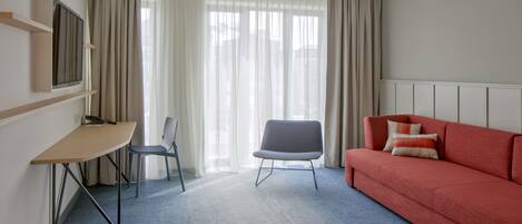 Una habitación de hotel con escritorio, silla, sofá rojo y un ventanal con cortinas.