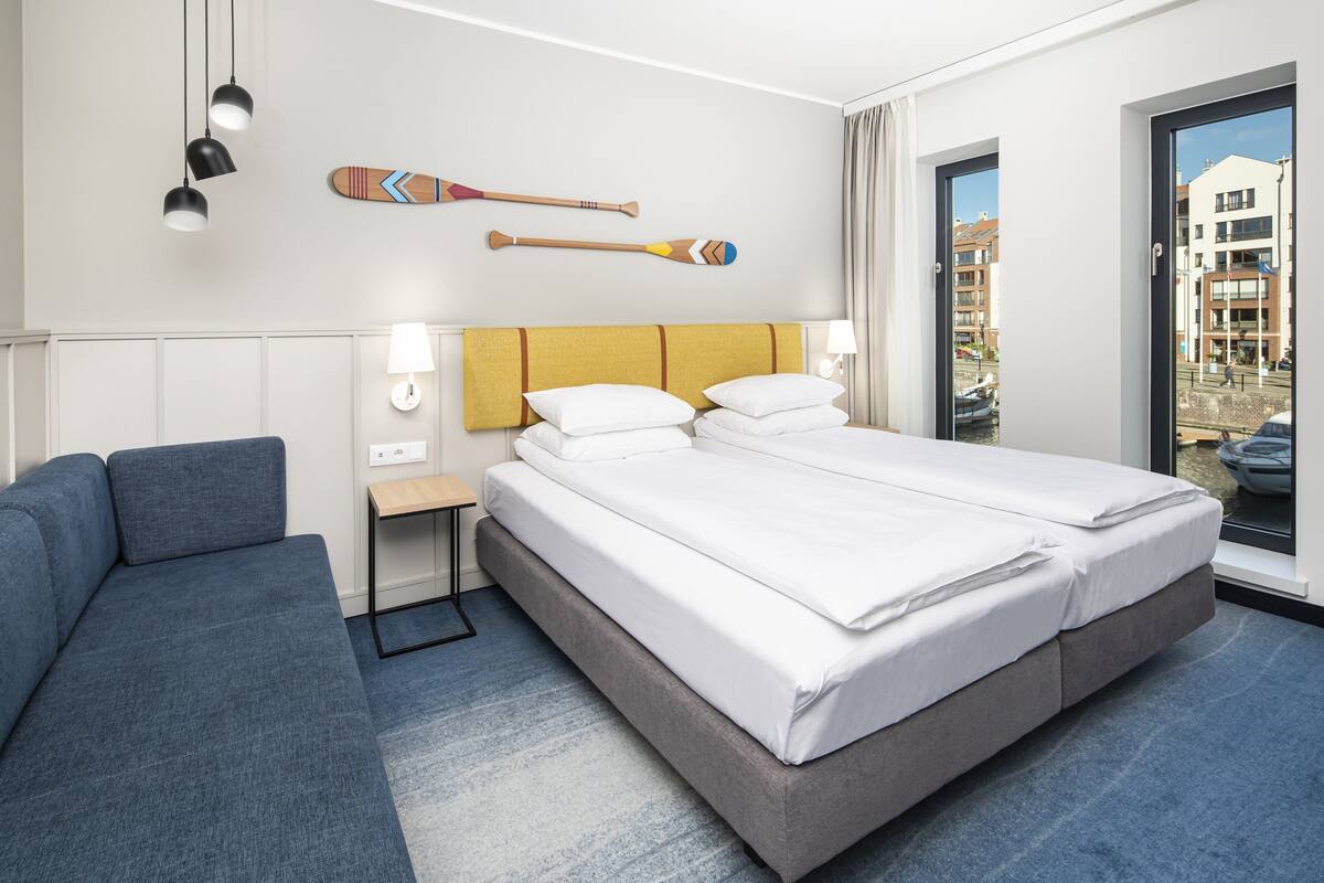 A hotel room with a double bed, a sofa, a small table, and a window with a view of buildings.
