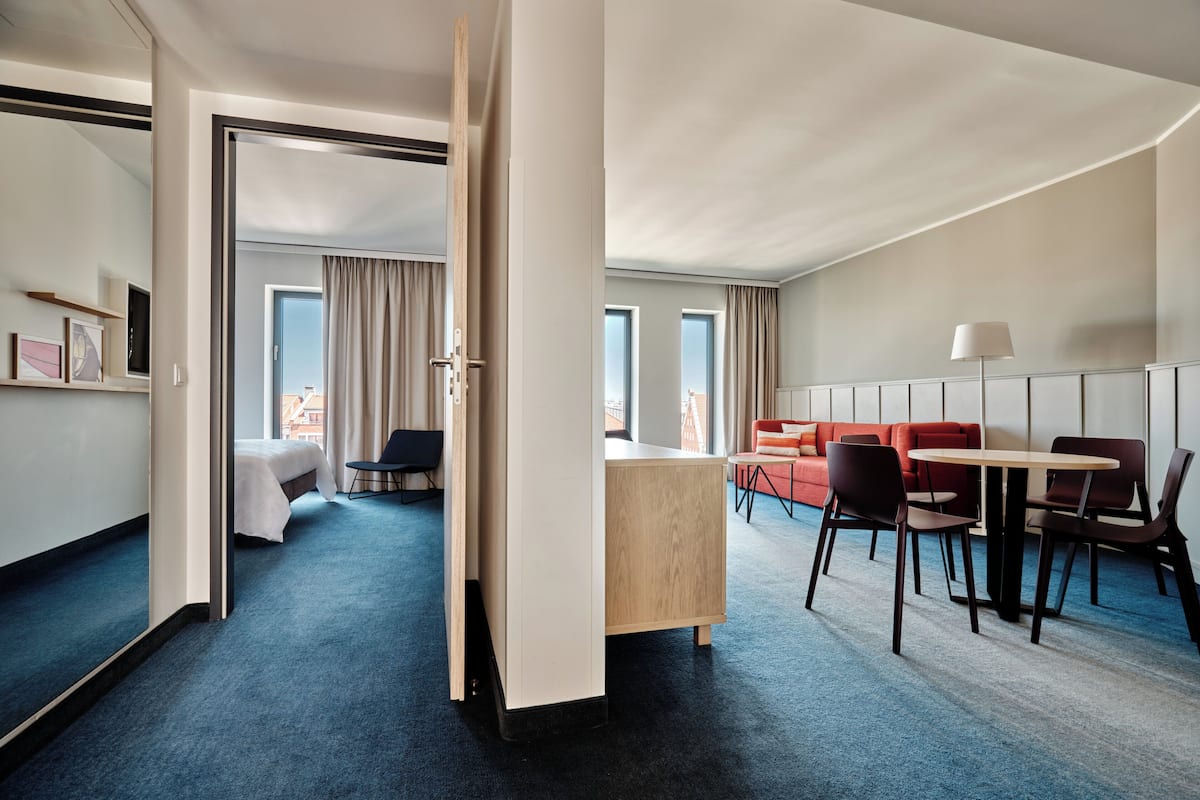 A hotel room with a bed, a desk, a dining area with chairs, and a sofa. There is a window with curtains, a door, and a view of the city.