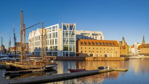 Uma marina com barcos atracados, um edifício moderno com grandes janelas e um edifício histórico de tijolos com uma torre do relógio ao fundo.