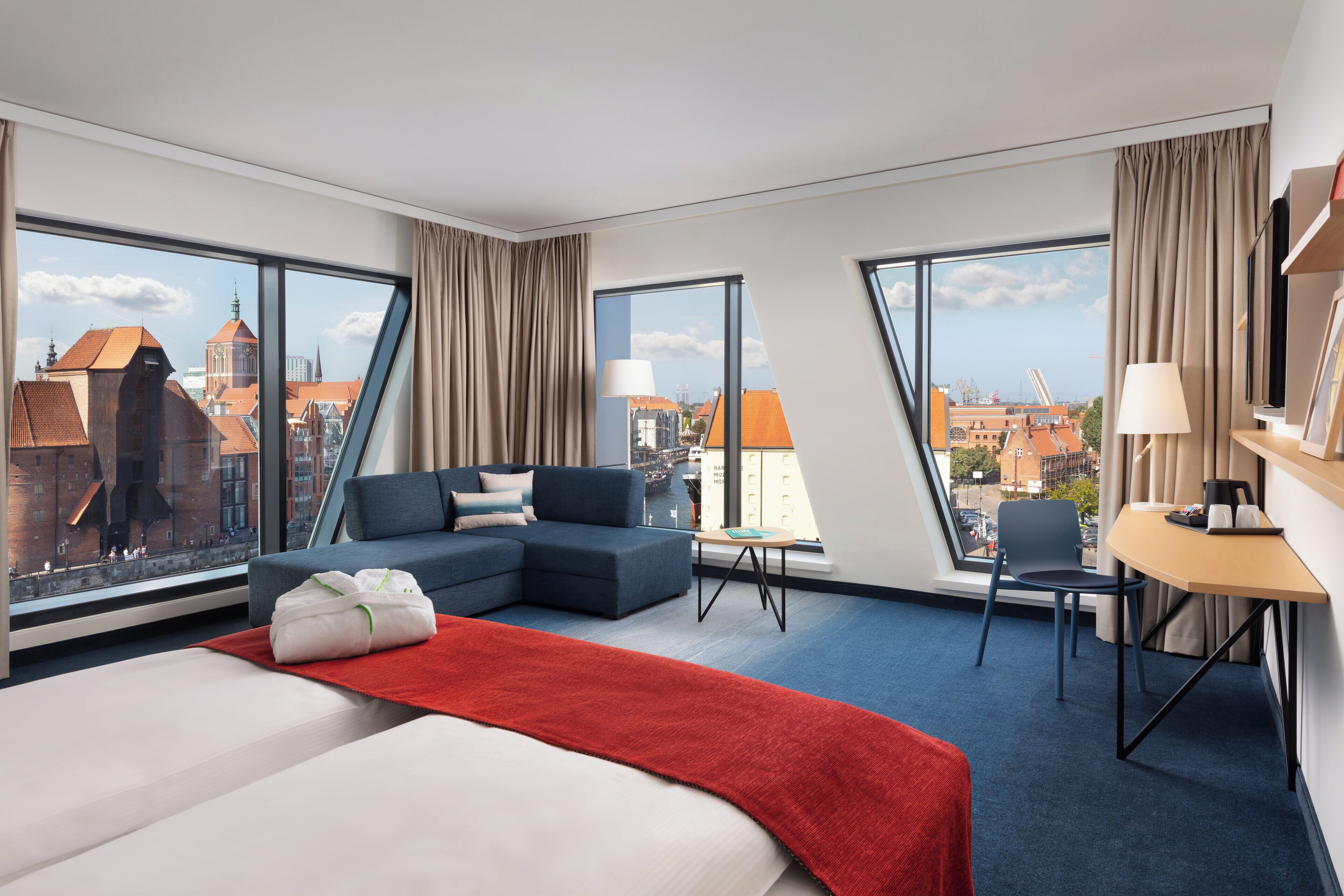 A modern hotel room with a large bed, a sofa, a desk, and large windows offering city views.