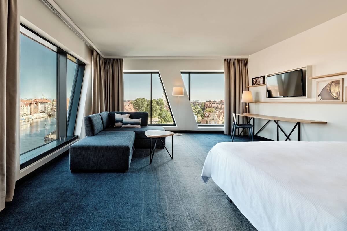 A modern hotel room with a large bed, a sofa, a desk, and a view of the city.