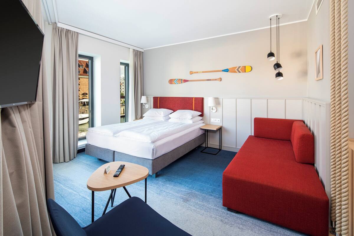 A hotel room with a bed, a red sofa, a small table, and a TV.