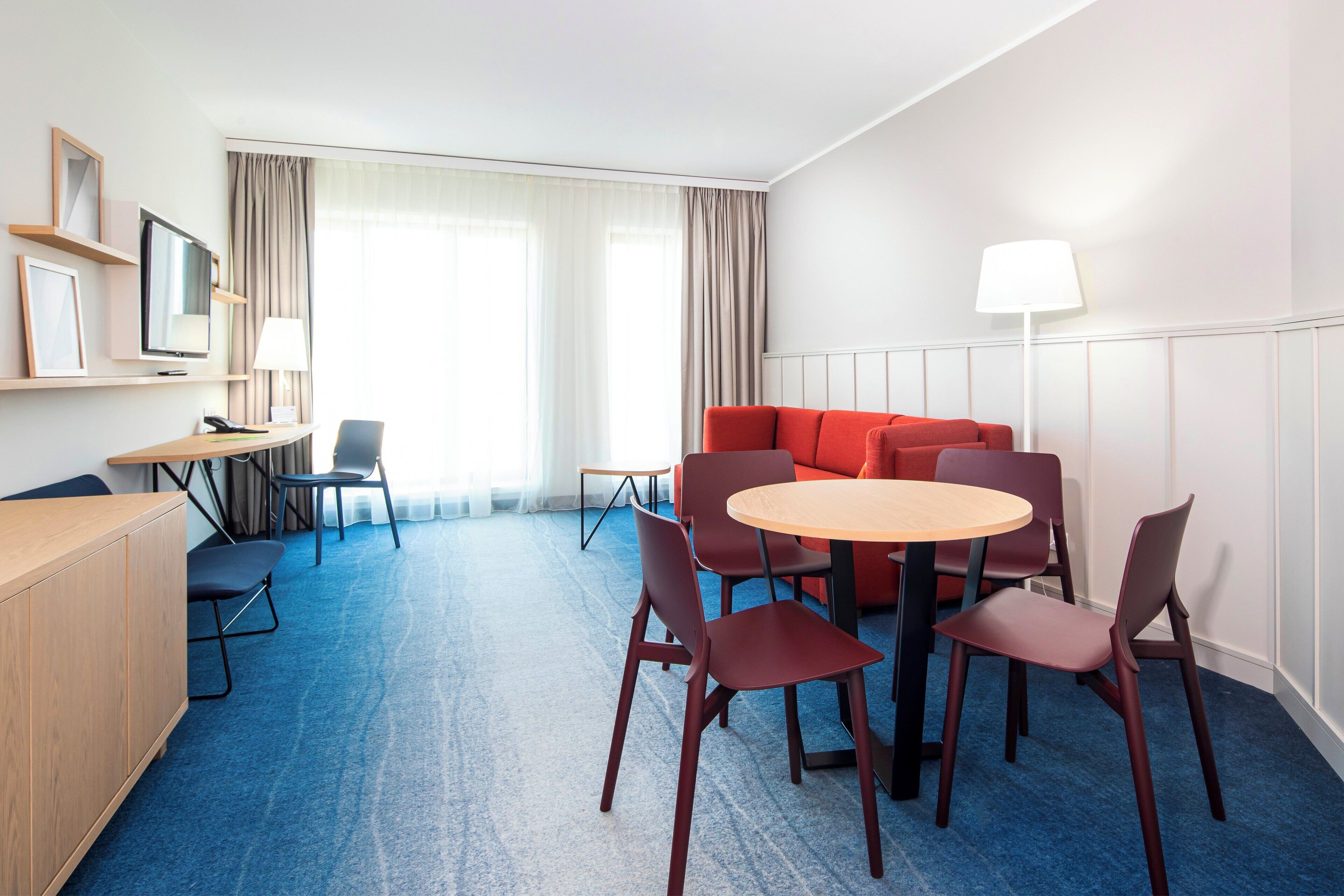A hotel room with a round table, four chairs, a sofa, a TV, a desk, and a lamp.