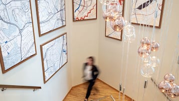 Un escalier avec des marches en bois et une rampe en verre, menant à un palier orné de cartes encadrées et sur lequel est suspendu un luminaire.