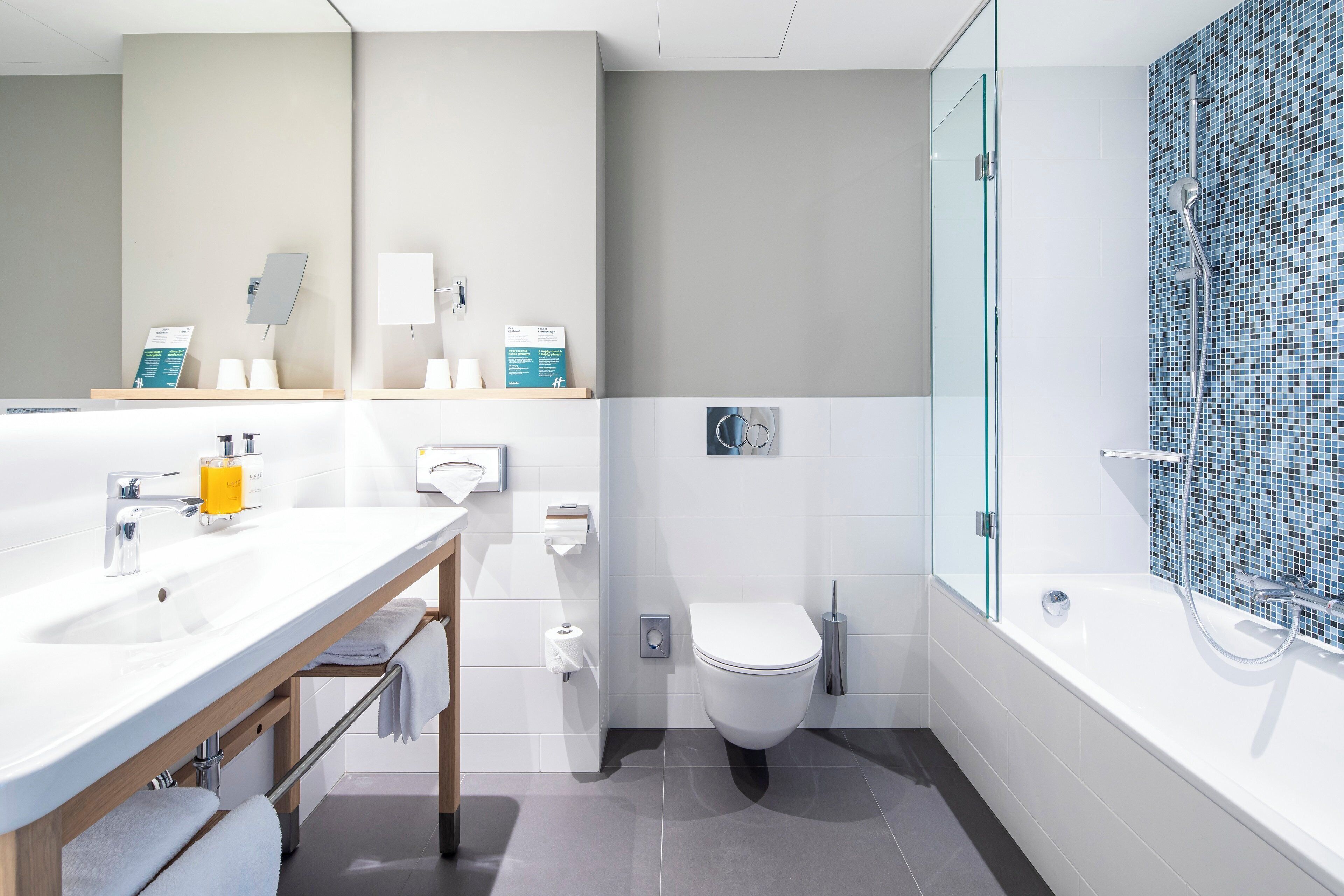Un baño moderno con lavamanos, inodoro y bañera blancos. Cuenta con puertas de cristal, ducha y pared con azulejos.