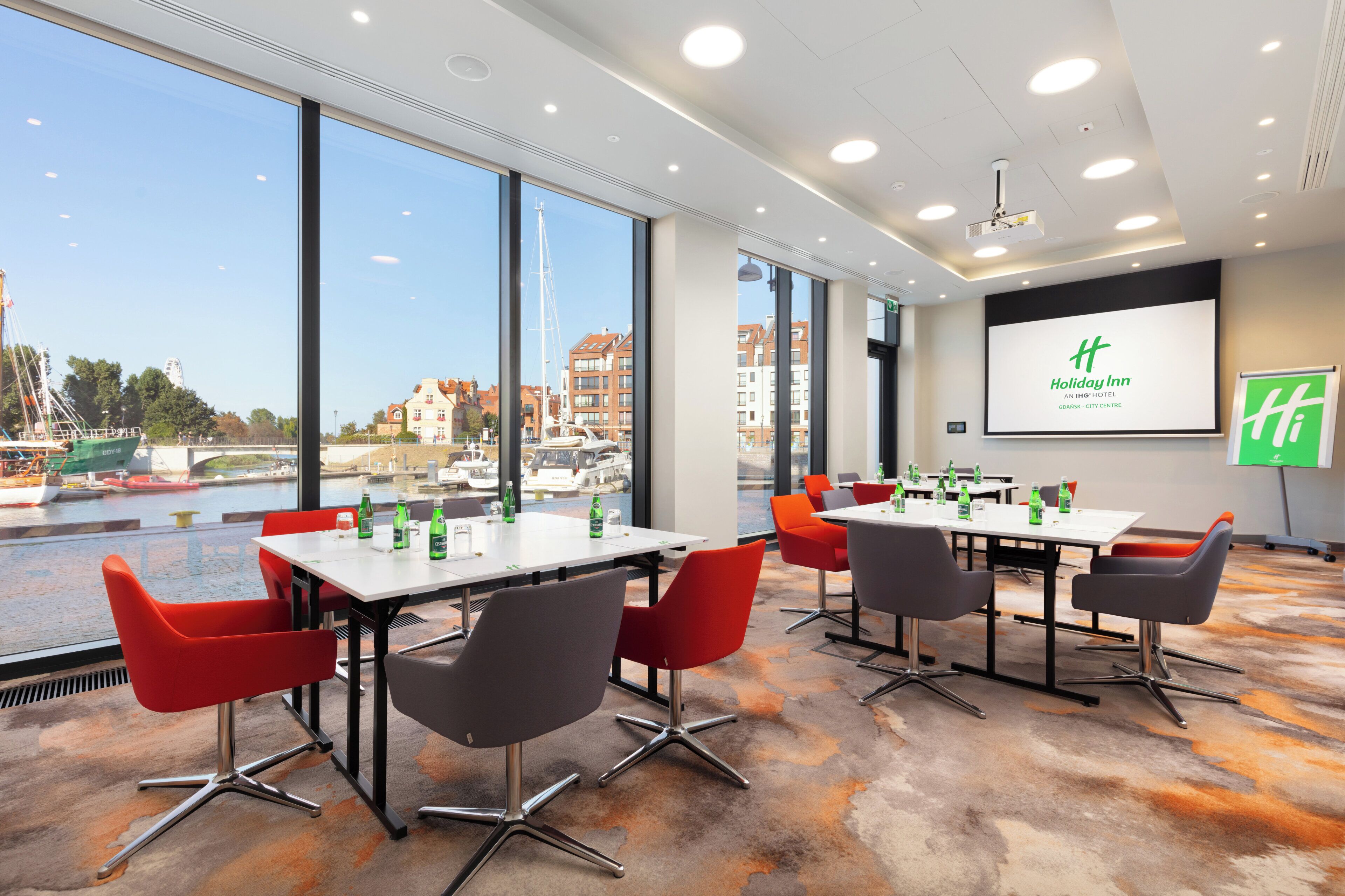 A conference room with large windows overlooking a marina, equipped with tables, chairs, and presentation screens.